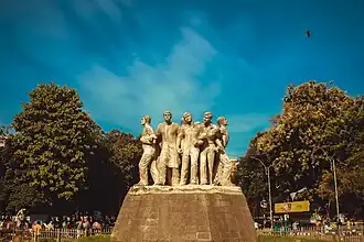 Four figures stand in a circle holding up the symbol of Wikipedia, which consists of a globe made of jigsaw pieces, with few pieces missing, symbolizing that the work of the encyclopedia will never be finished