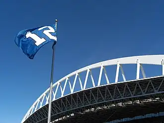 A blue flag with a white number 12 flies against a clear sky. An expansive white roof truss is behind the flagpole.