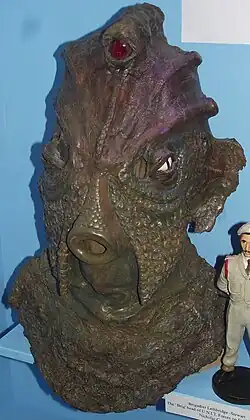 The head of a reptilian creature, with brown skin, a rounded mouth, and two lizard-like eyes, with a third red eye at the top of its head
