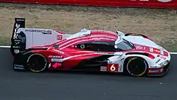 Colour photograph of a white, red, and black-liveried Porsche 963 hypercar at Le Mans, shot from the side and slightly above.