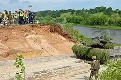 A tank driving out of water, people around watching