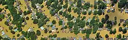About 100 colorful roofs and green trees completely surrounded by tan flood water.