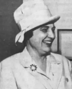 A smiling white woman with dark hair, wearing a white brimmed hat with a high crown, and a white suit