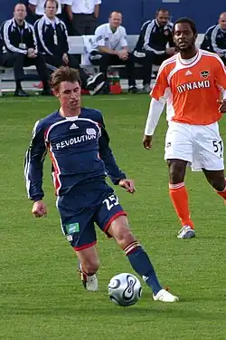 A soccer player in a New England Revolution jersey dribbles the ball, while another one in a Houston Dynamo jersey stands behind him.