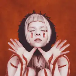 Aurora is against a reddish background with her eyes closed and thumbs under her chin while her palms are open. She has red marks on her skin and a black scarf around her neck.