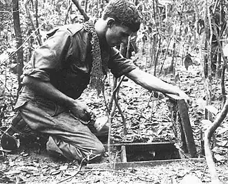 A soldier is kneeling over an open trapdoor on the jungle floor while holding a torch.