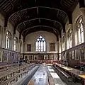 Interior, Hall, Balliol College, Oxford (1883) as with his hall at Girton College, the interior relies on the raw building materials there is no attempt to use tile work or other elaborate decoration save simple wood panelling and natural stonework, note the typically solid wooden braces in the roof resting on corbels that have coats of arms on them, there is also simple armorial stained glass in the end window.