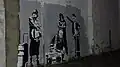 A gray wall is painted with black and white images. A 1980s style boom box sits on the ground. Older adults are dressed in 1980s street style clothing.