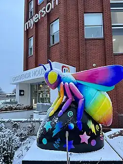 A brightly coloured bee sculpture covered lightly in snow, positioned outside the entrance of Grosvenor House, home to Myerson Solicitors in Altrincham. The sculpture features rainbow wings and limbs with a black base decorated with stars, lightning bolts, and other vibrant patterns. Snow blankets the surrounding bushes and ground, with the red-brick office building in the background.