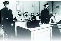Black and white photograph of Menahem Mendel Beilis at the trial with two guard on the side and other people behind him.