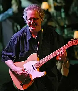 A man wearing glasses, playing a guitar and standing behind a cymbal.