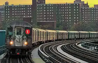 A 1 train, composed of R62A cars, is seen above ground entering the 207th Street station. The front of the train contains two white lights providing slight illumination, two windows, a door, and the Symbol for the 1 line on the left window.
