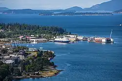 The waterfront on the left side with multiple large ships and small sail boats in the water. Mountains are visible in the distance