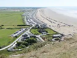 Looking due north at a beach, caravan sites and village in the distance seen from the top of a hill