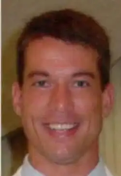 A smiling, clean-shaven young man with short dark hair wearing a white shirt and silvery tie against a blue background