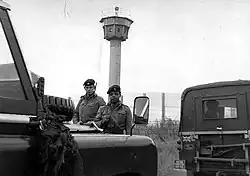 Two British soldiers carrying rifles standing behind a pair of Land-Rover vehicles, one of which has a "British Frontier Service" plate. Behind them is a high mesh fence, behind which is a tall watchtower with an octagonal cabin at its top.