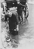 An elderly woman gathers vegetable waste tossed from a vegetable seller's wagon for her lunch, 1923