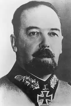 The head of a man with beard, shown in semi-profile. He wears a military uniform with a military decoration in shape of an iron cross displayed at the front of his shirt collar. His hair is dark and combed to back.