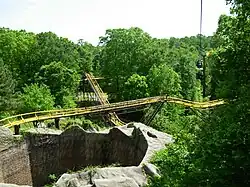 The Loch Ness Monster's second lift hill and mid-course brake run is viewed in the image among green foliage and trees. The mid-course brake run is above the second lift hill in the center image. The tunnel's exterior structure can also be seen in the bottom left.
