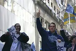 A black man on the left wearing a white T-shirt and dark jacket smiles holding a sign, while a white man on the right wearing a blue sweatshirt which reads "Champions" points upwards. White streamers hang from a building in the background.