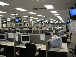 A large room with a suspended ceiling packed with cubicles containing computer monitors