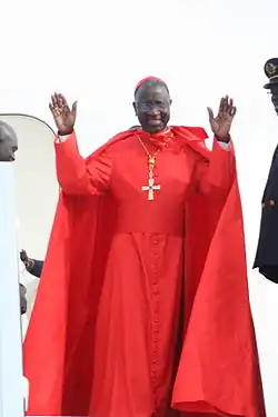 A Black man wearing a red cassock and cap with his hands raised.
