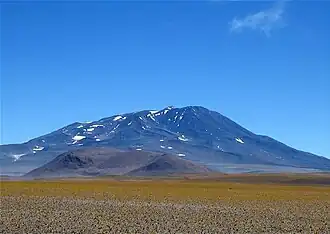 A mountain with snow patches rises above a smaller hill, which in turn rises above a plain covered with sparse yellow plants