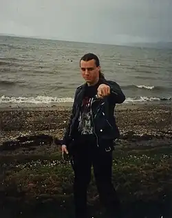 A man, Chuck Schuldiner, is shown on a dark shoreline. He has long hair, black pants and a black shirt, and a black leather jacket.