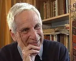 Colour photograph of a clean-shaven, smiling, white-haired man, resting his chin on his hand, with book shelves in the background