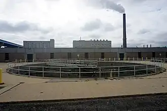 A circular holding tank in the foreground with buildings and a smokestack in the background