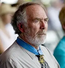 Profile of a white man with a full, gray beard wearing a star-shaped medal from a blue ribbon around his neck.