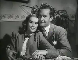 Black and white movie still showing Sally Ann Howes and Griffith Jones, both in fine suits, smiling and leaning against each other
