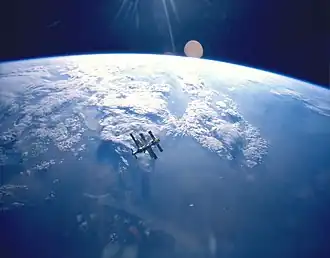 A cluster of modules and feathery solar arrays floats in the middle distance before an image of the Earth and the blackness of space above its horizon. Sunrays project from the top centre of the image.