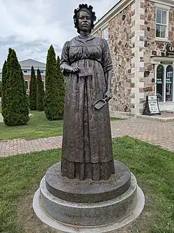 Statue on a short stone pedestal of a Black woman in a long dress and bonnet, holding a scoop in one hand and a slaip of paper in the other