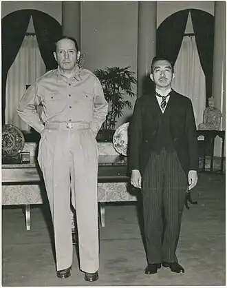 A tall Caucasian male (MacArthur), without hat and wearing open-necked shirt and trousers, standing beside a much shorter Asian man (Hirohito) in a dark suit.
