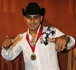 A dark man with black hat, white shirt, raising both hands, with a medal with red ribbon around his neck and legend BMI in the front.