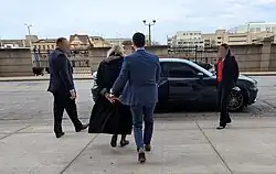 A man in a blue suit escorts Hannah Dugan, who is wearing a pair of handcuffs, to a black van. Two individuals, one male and one female, with blurred faces stand on each end of the vehicle.