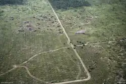 Aerial photo of a large open area bordered by thick vegetation. A road runs from top to bottom through the middle of the position while a range of field defences, earthworks and armoured vehicles are evident. Smoke is rising from the position.