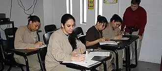 Four female prisoners in beige uniforms seated at desks . A teacher is supervising one of them.