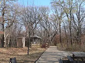 Boardwalk running through woods in fall; wooden picnic table and wood display structures beside trail