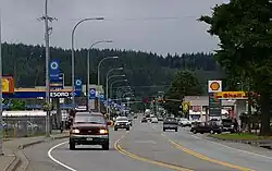 Photo of a three-lane main street in a small town. Visible is a traffic light, sparse traffic, and businesses such as a car lot and gas station.