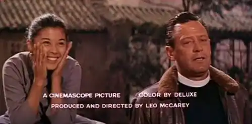 An East Asian young woman smiling with her hands on her cheeks and a white priest in a pilot jacket. A wooden building is in the background. In the foreground, the text "A CINEMASCOPE PICTURE COLOR BY DELUXE / PRODUCED AND DIRECTED BY LEO MCCAREY