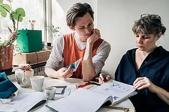 Two people sitting at a table by a sunny window looking together at fashion sketches and enjoying tea.