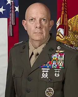 Official Marine Corps portrait of General David H. Berger - caucasian bald male with dark blue eyes, wearing Marine Corps service uniform - olive-colored jacket with shoulder epaulets with 4 silver stars, left chests covered with military ribbons, partial USA on flag showing on left, partial Marine Corps flag showing on right