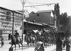 Soldiers on horseback trot along a road past several buildings watched by a group of civilians