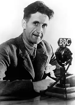 A man with a thin mustache in a thick suit sitting in front of an old-style microphone which reads "BBC"