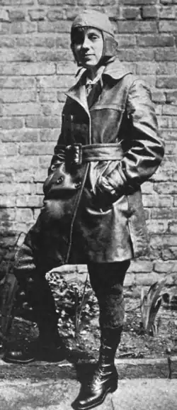 A young white woman dressed in a leather cap and thigh-lenght coat, wearing lettings and boots, standing in front of a brick wall with one leg bent at the knee