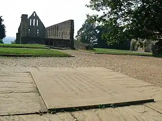 Photograph shot low to the ground near an inscribed stone set into the ground, with a background of ruined stone buildings.