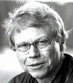 Black and white publicity shot of a half-smiling, clean-shaven, bespectacled man, with slightly windblown blond hair. His features are not unlike the musician John Denver.