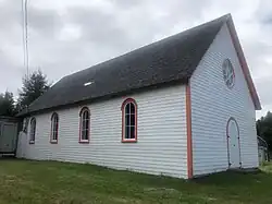 Photo of a single storey wooden building, painted white with orange trim. It has a circular window and an arched door in the gable end, and arched windows along the side facing the viewer.